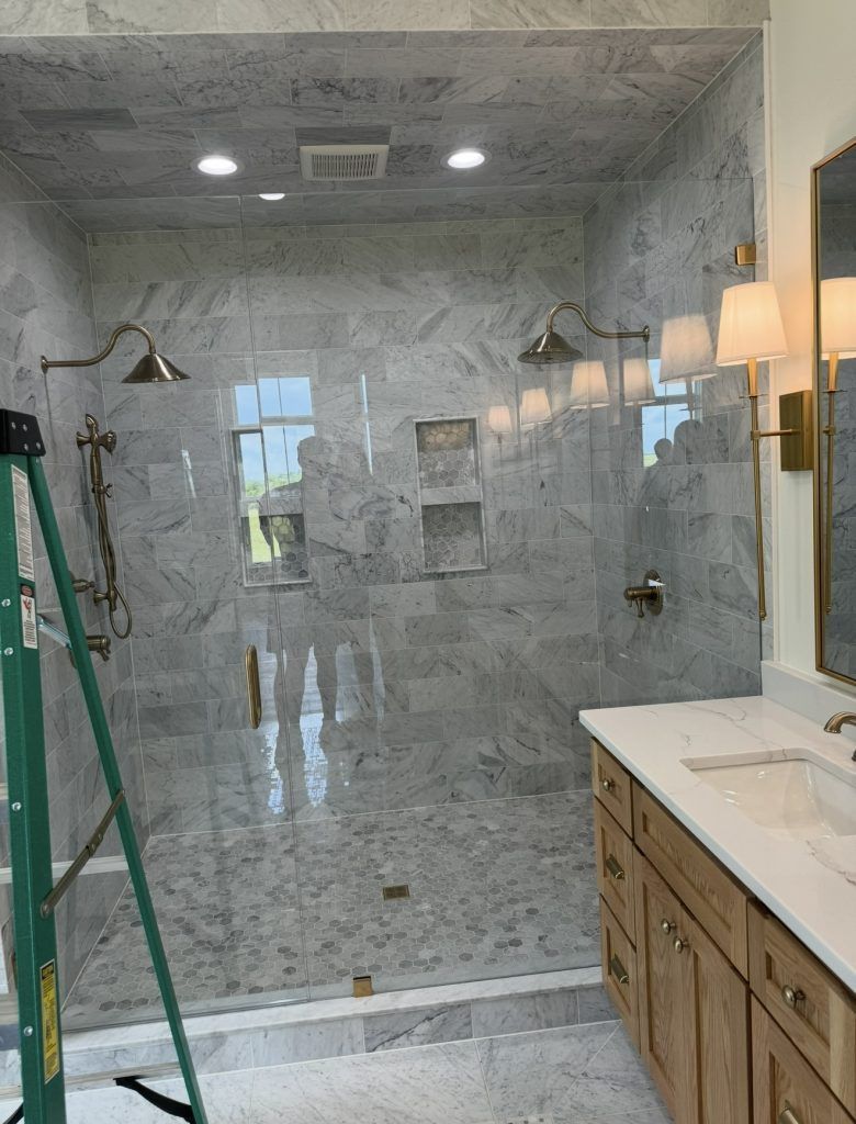 A marble-tiled walk-in shower with dual showerheads and a glass door beside a wooden vanity in a modern bathroom.