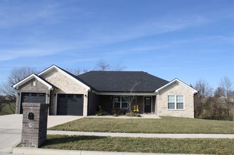 A tan brick single-story house with a black roof and attached garage sits behind a manicured lawn under a blue sky.