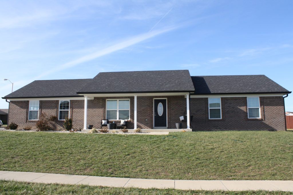 Single-story brown brick house with a dark gabled roof and a small covered porch under a bright blue sky.