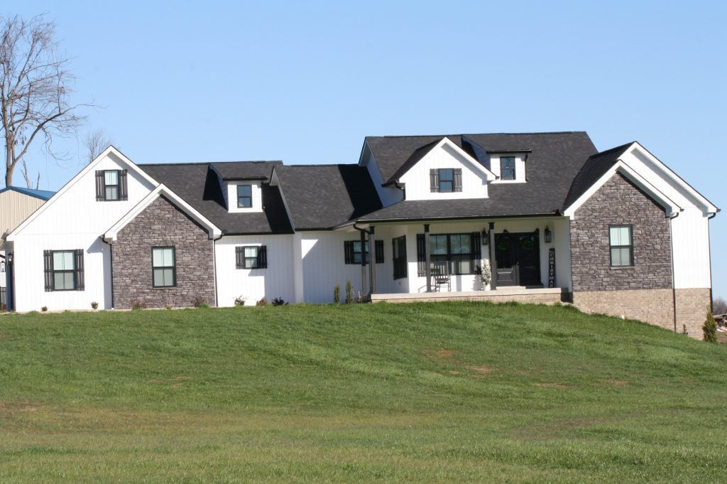 A modern farmhouse with white siding, dark stone accents, and a black shingled roof, set atop a grassy hill.