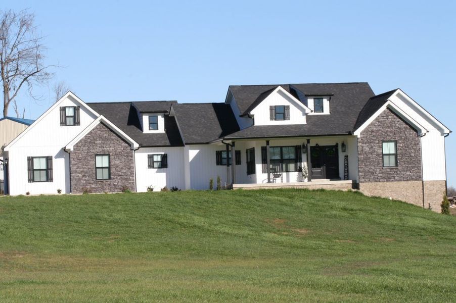 A white, multi-gabled house with dark stone accents and a dark roof, situated on a grassy hill under a clear blue sky.