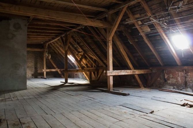 Empty attic with wooden beams, slanted roof, and a bright window light on worn floorboards.