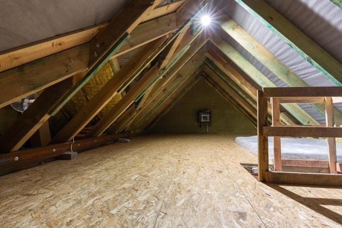 Attic with exposed wooden rafters and OSB floor, lit by a bright overhead work light.