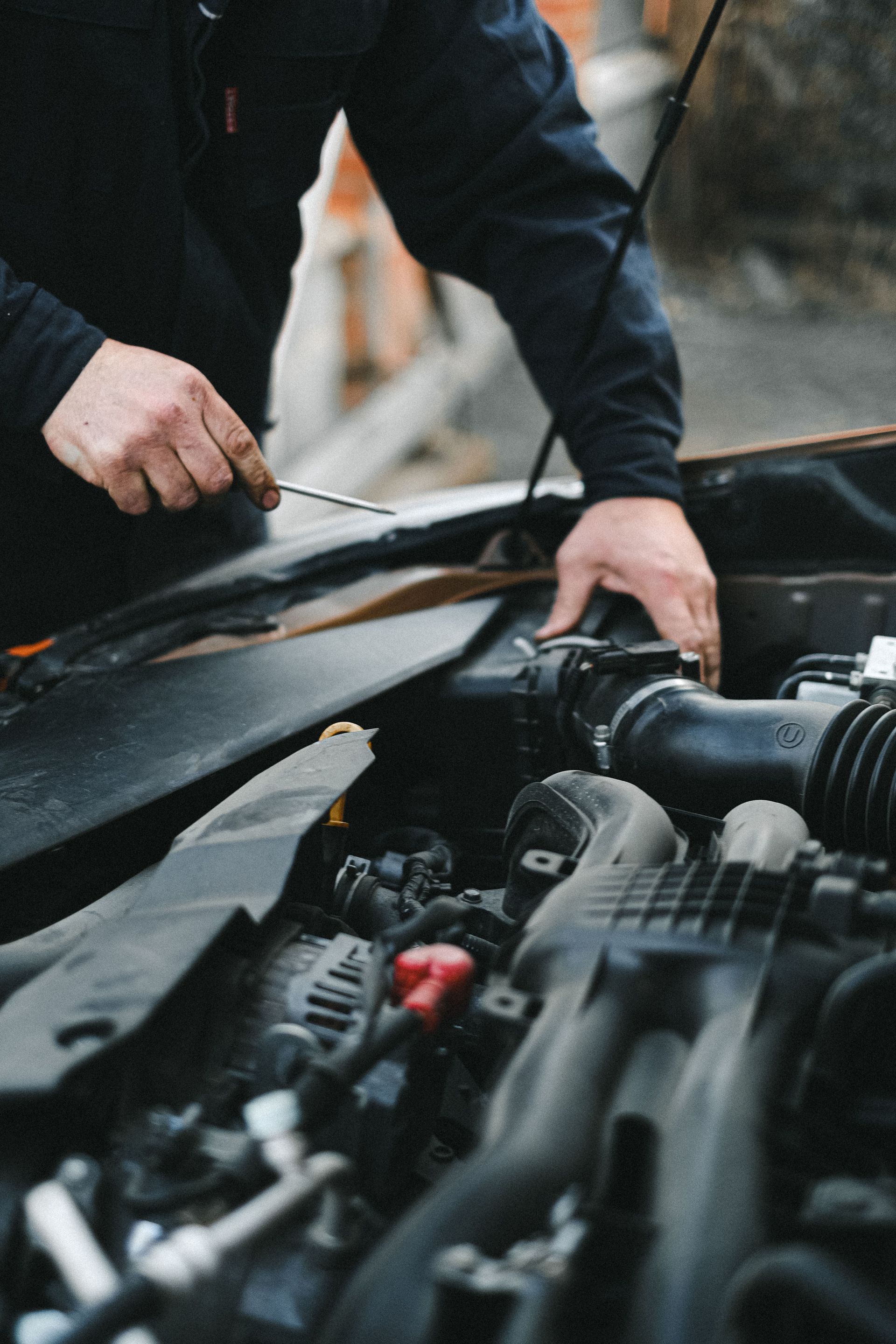 Mechanic working under an open car hood outdoors, hands inspecting the engine.