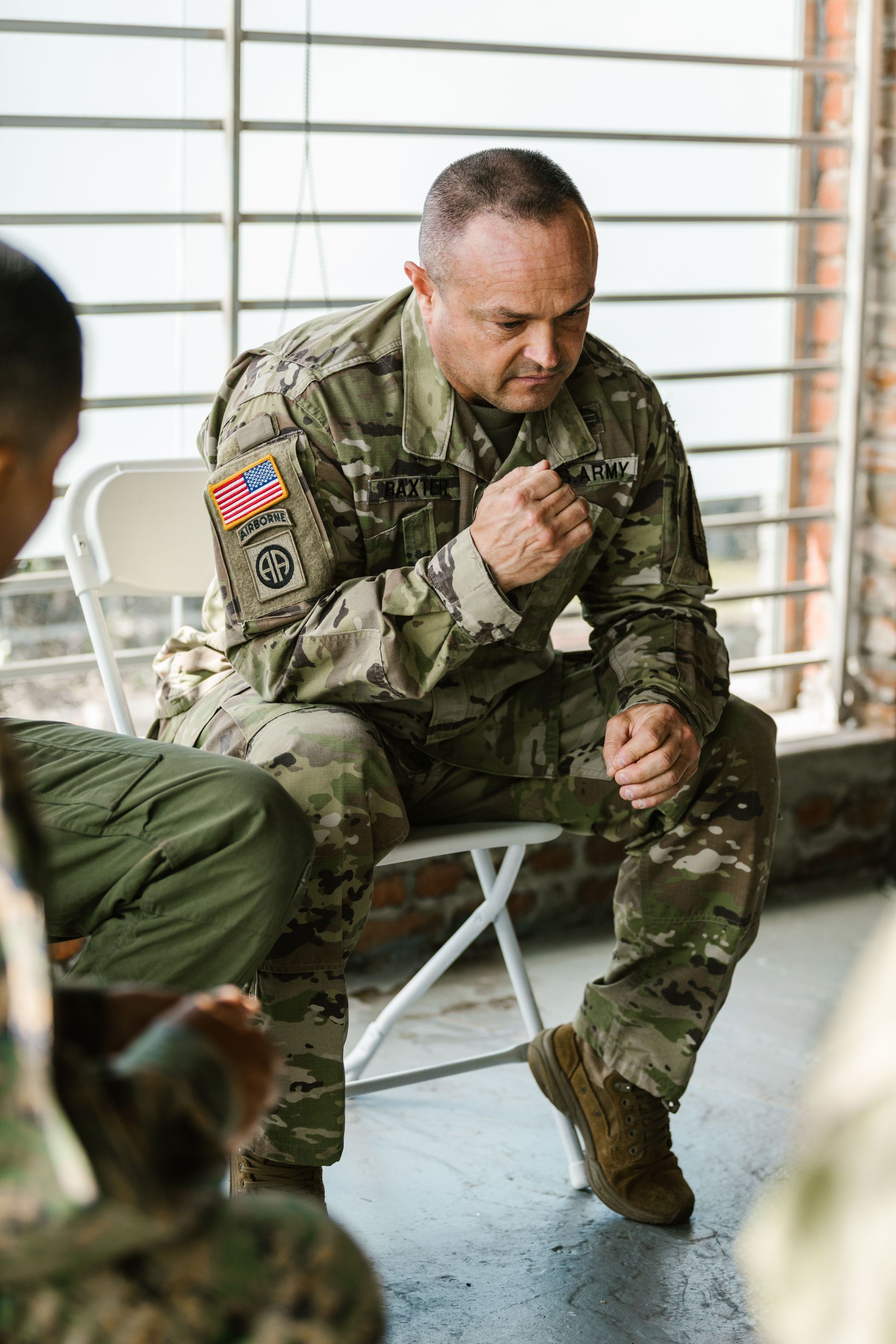 Soldier in camouflage uniform seated indoors, speaking while holding a microphone