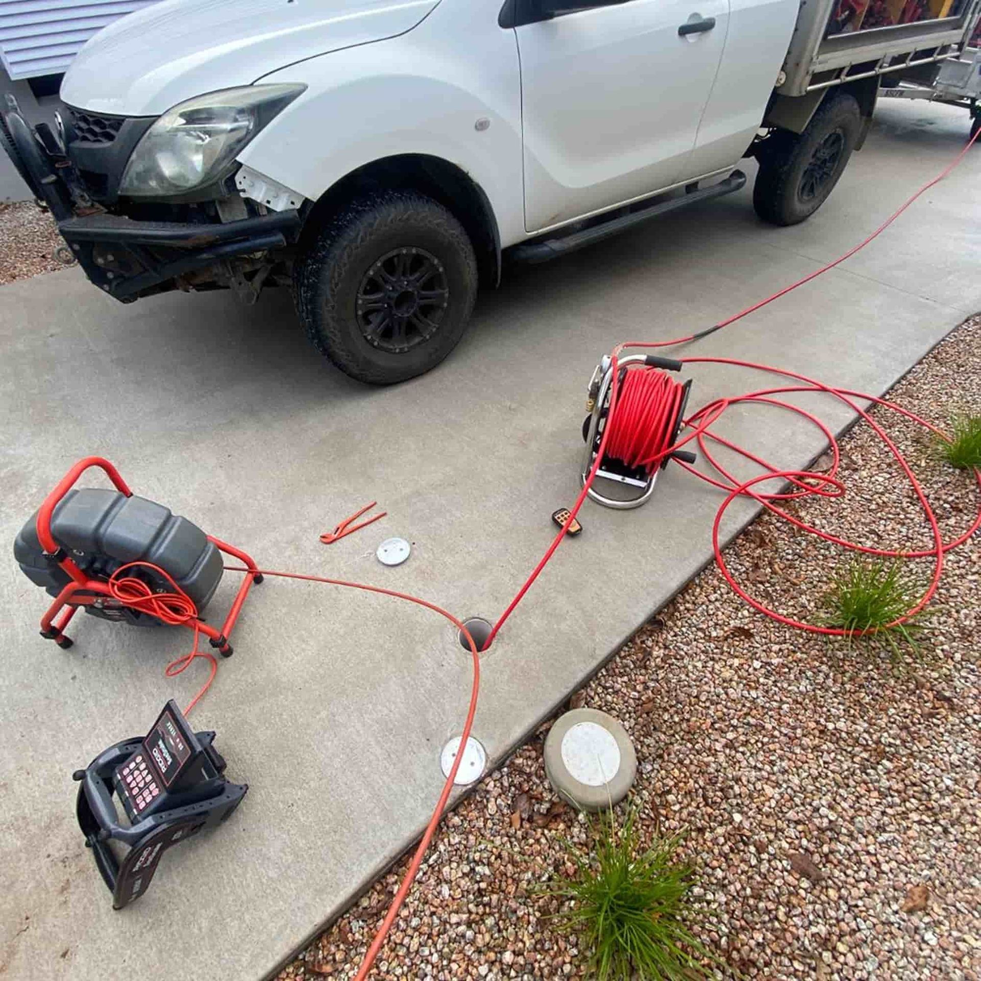 White truck with plumbing equipment on driveway; red cable connected to the truck and a camera monitor.