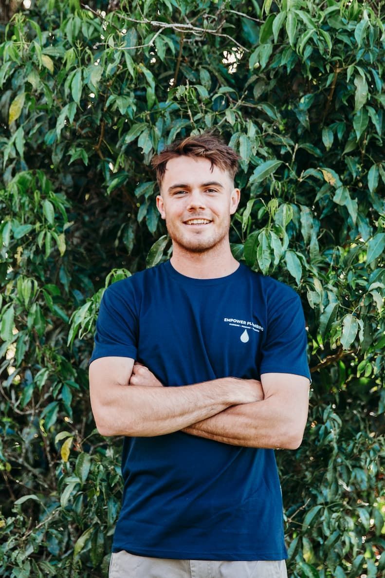 Man with arms crossed, smiling, wearing a blue shirt with a logo, standing in front of green foliage.