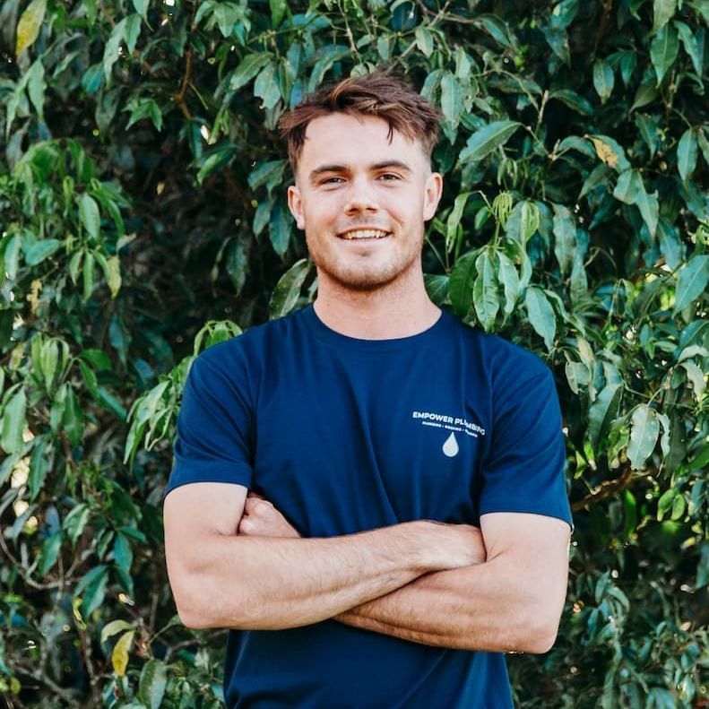 Man in blue shirt, arms crossed, smiling in front of green foliage.