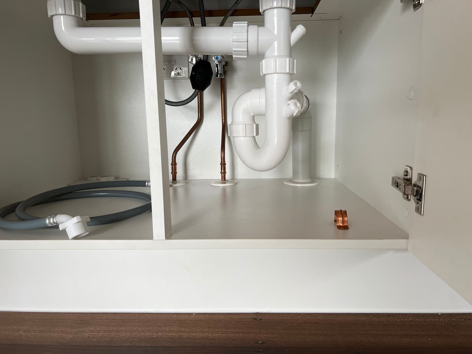 Under-sink cabinet with plumbing; white pipes and a drain, copper pipes, and a grey hose against a white shelf.
