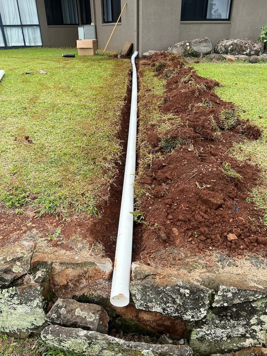 A white PVC pipe runs through a dug trench in a grassy yard, next to a rock wall.