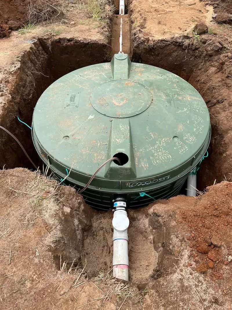 Green septic tank in an excavated hole, with connected pipes and a lid.