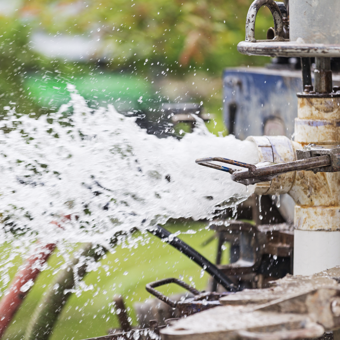 Water gushing from well drilling equipment, with droplets spraying in the air; green grass and a machine in the background.