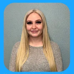 Woman with long blonde hair smiles, wearing a gray sweater, standing in front of a blue background.