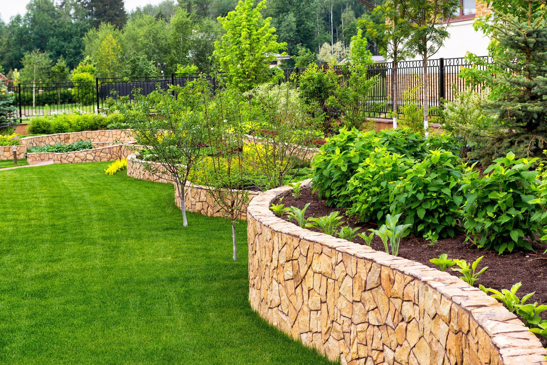 Curved stone garden wall bordering a lush green lawn and landscaped shrubs with trees in the background