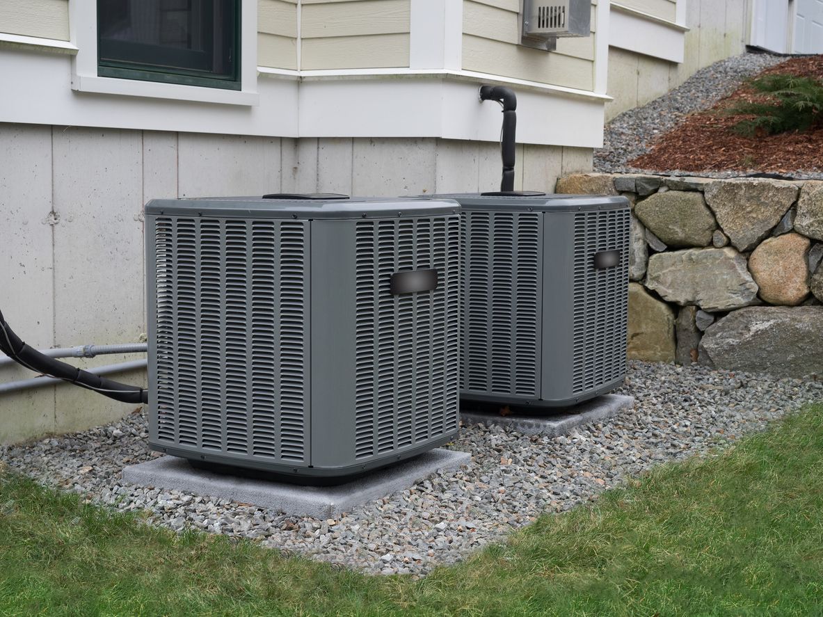 Two gray outdoor HVAC units beside a house on a gravel bed
