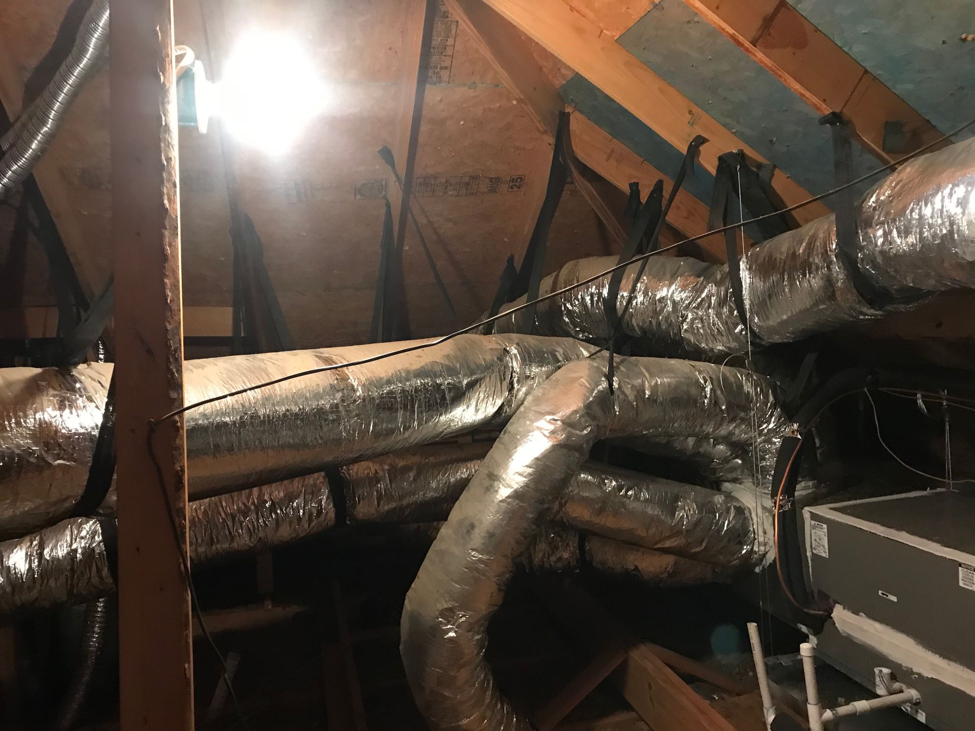 Attic with exposed ductwork and insulation under sloped rafters, lit by a bright bulb.