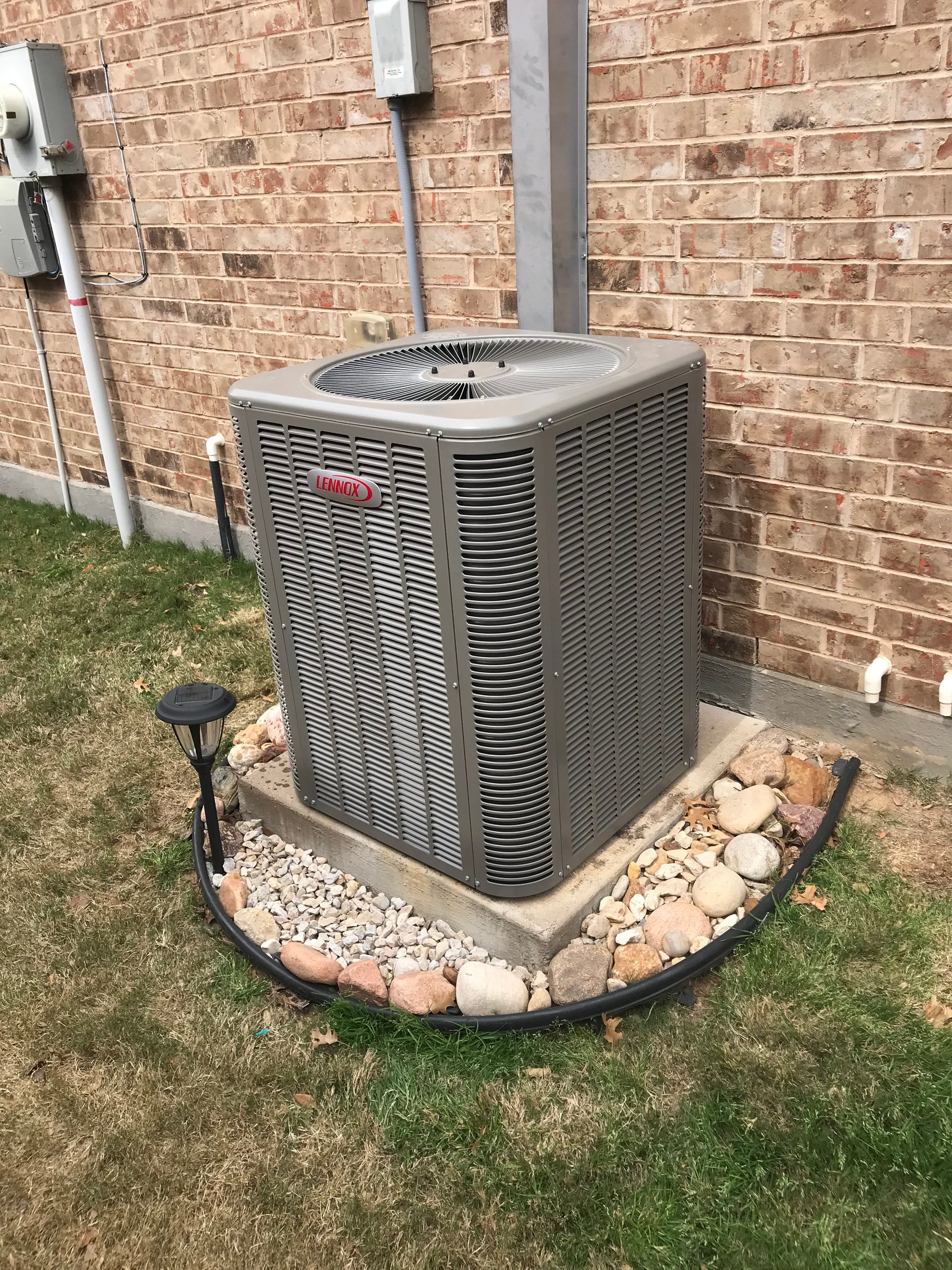 Outdoor air conditioner unit beside a brick house wall, surrounded by a stone border and grass.
