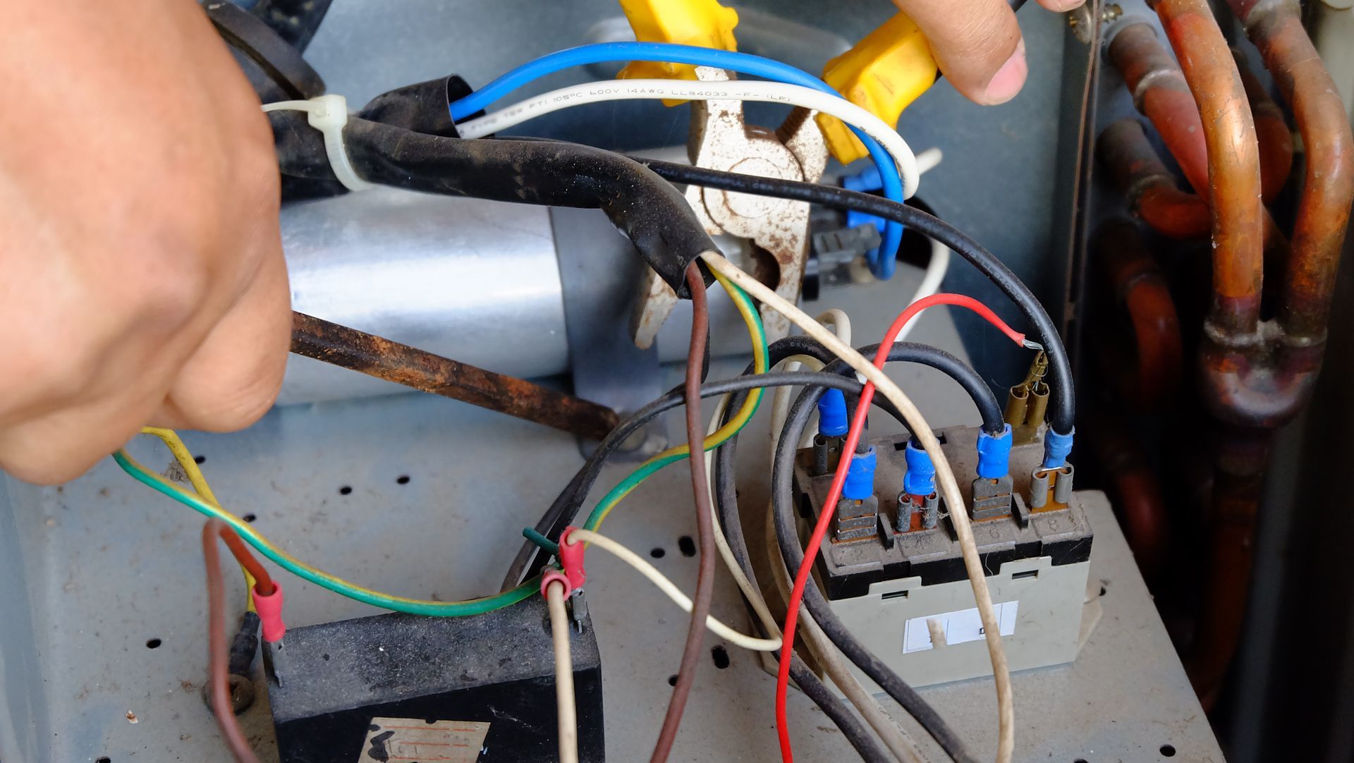 Hand connecting loose wires inside an electrical unit with gray panels and copper pipes
