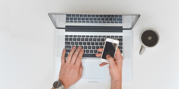 Hands typing on a laptop while holding a smartphone beside a coffee cup on a white desk
