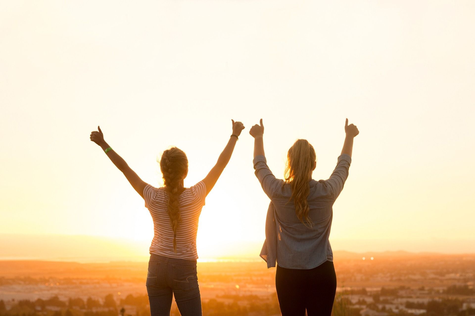 Resilienz: Deine innere Stärke für ein erfülltes Leben Zwei Frauen, die Ihre Hände zum Himmel strecken. Die Daumen zeigen nach oben. Während sie die Aussicht von oben auf eine Stadt genießen.