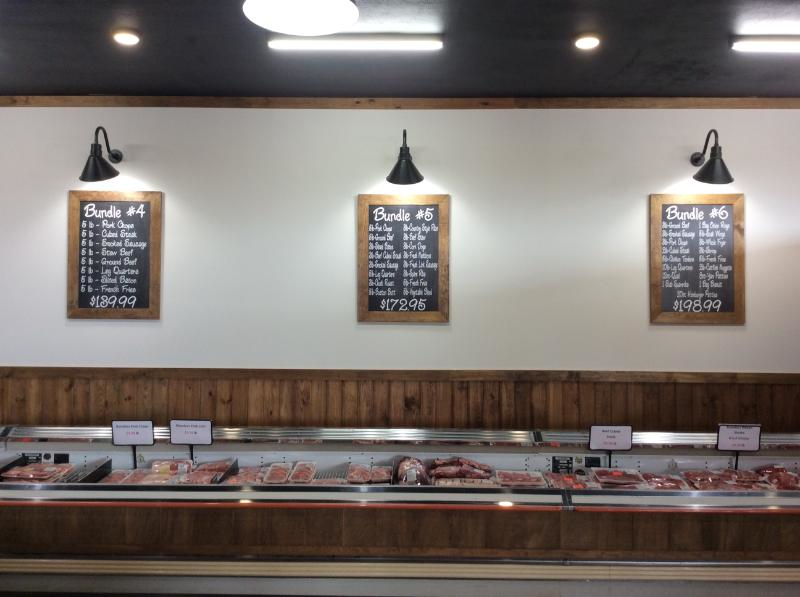 A butcher shop counter featuring three chalkboard menus listing meat bundles on a white wall above refrigerated cases.