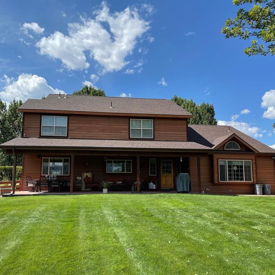 A two-story brown house with a large covered porch and a mowed lawn under a sunny blue sky with clouds.