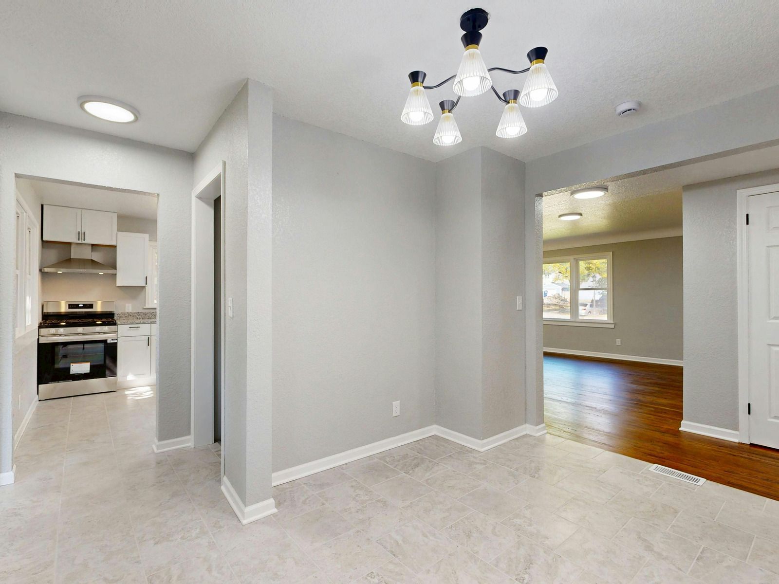 A bright dining area features light tile floors, a modern chandelier, and an open view into a kitchen and living space.