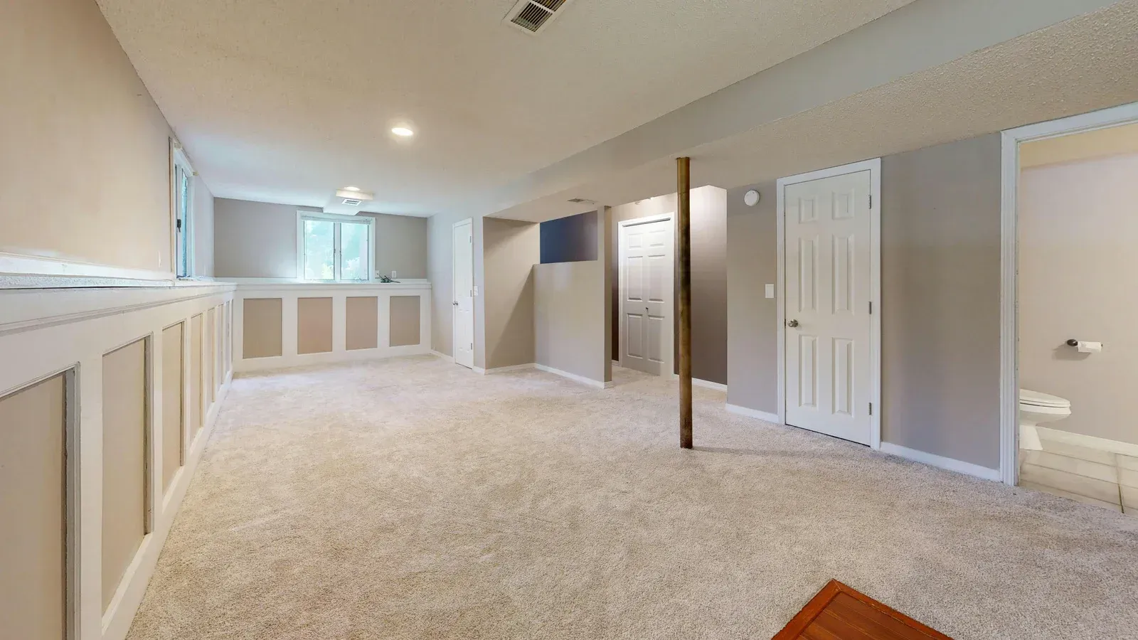 A neutral-toned, carpeted basement with wainscoting, a support column, two closed white doors, and an open bathroom entry.