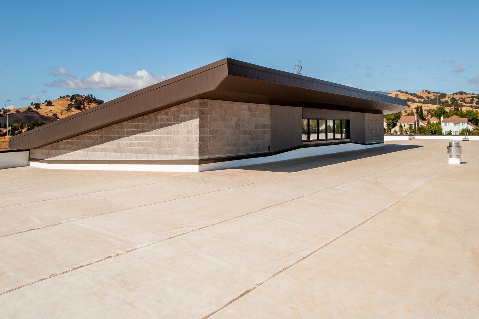 A view of a tan flat roof featuring a low-profile architectural structure with a sloping brown roof and stone-textured wall.