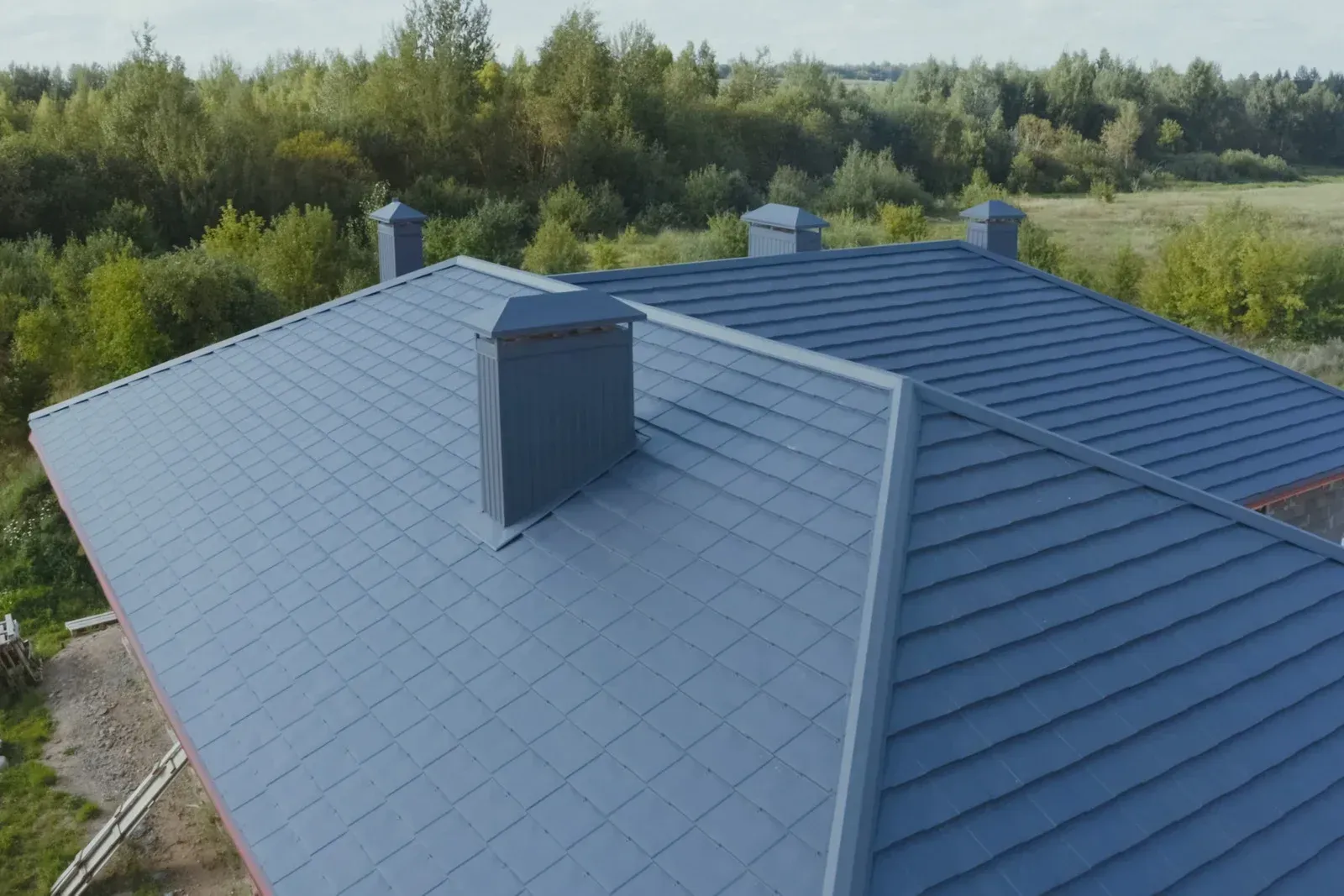 An aerial view of a dark grey tiled hip roof with three chimneys, surrounded by green trees and a grassy field.