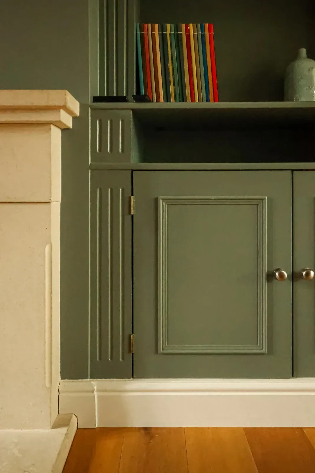 A sage green built-in bookshelf with cabinets beside a light stone fireplace, featuring wooden floors and white trim.
