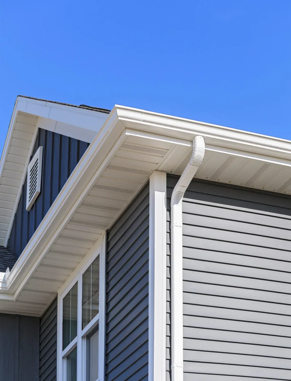 Close-up of a house exterior showing gray siding, white soffit, fascia, corner trim, and a white downspout against blue sky.