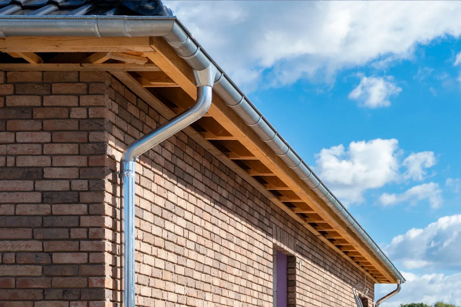 A metal gutter and downspout system installed on the brick wall and wooden roof eaves of a house under a blue sky.