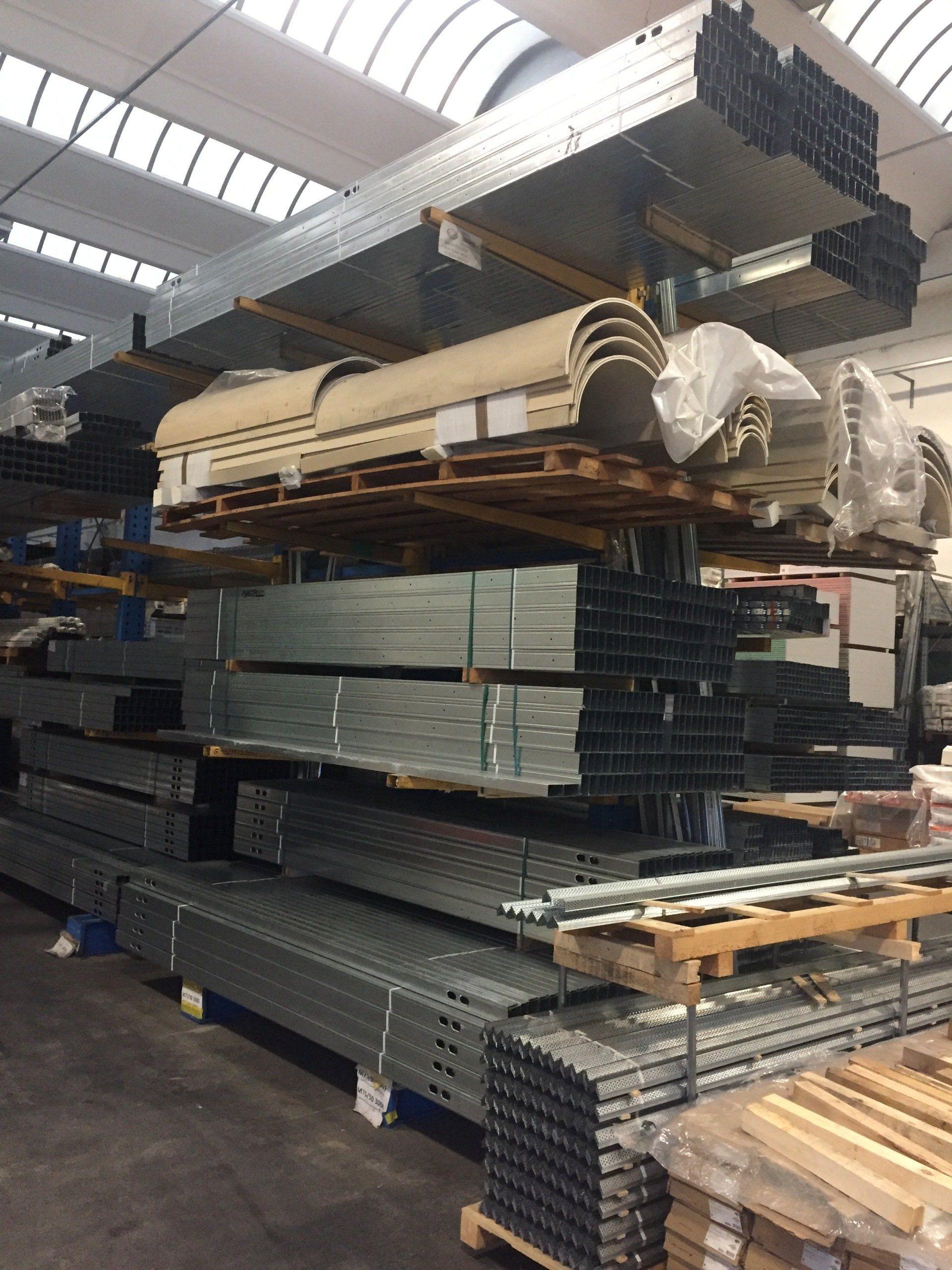 Tubi metallici impilati e legname stoccati su scaffalature da magazzino sotto un tetto con lucernari.