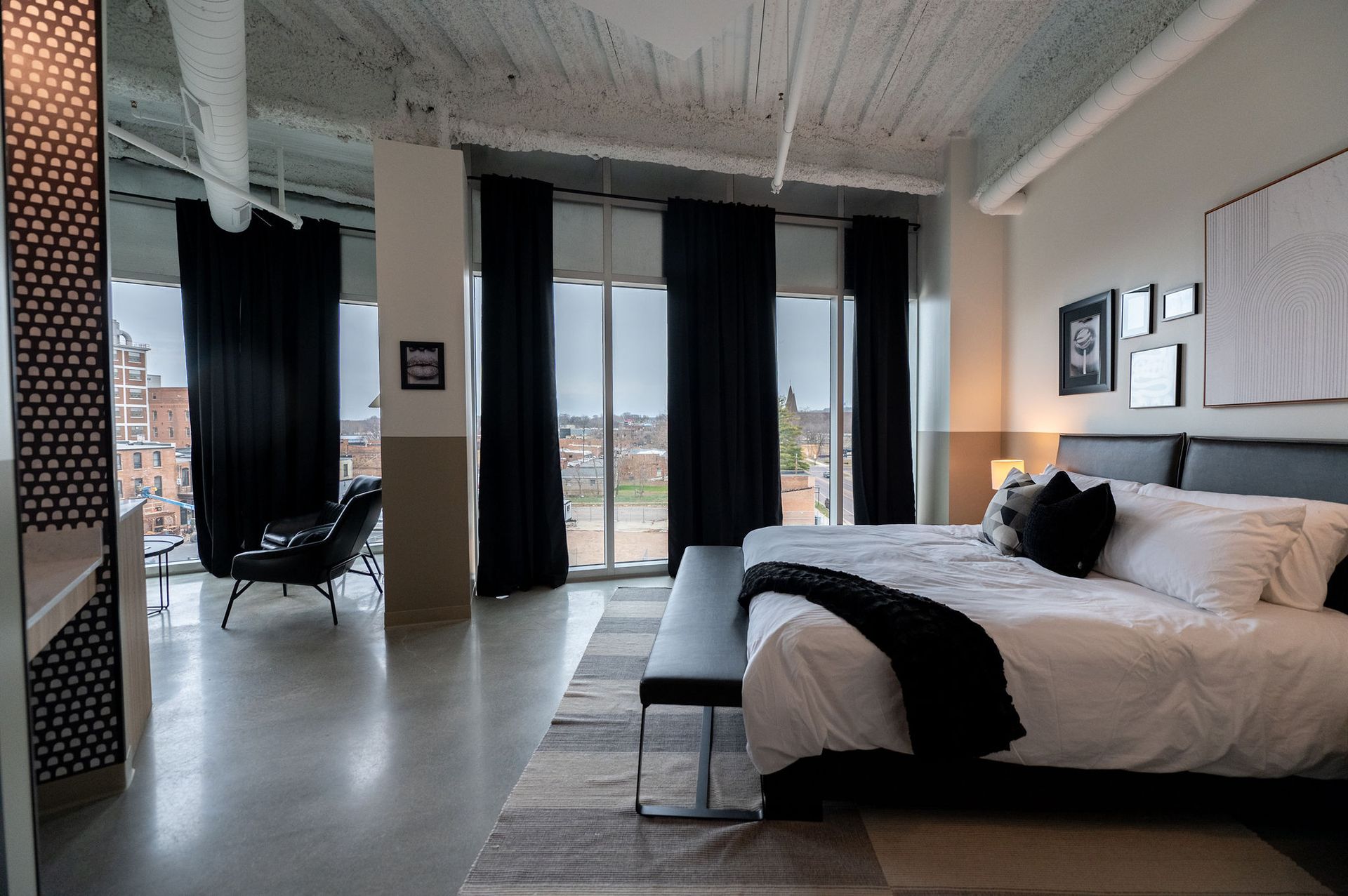 Modern bedroom with large windows, dark curtains, and a bed with white bedding in a loft-style apartment