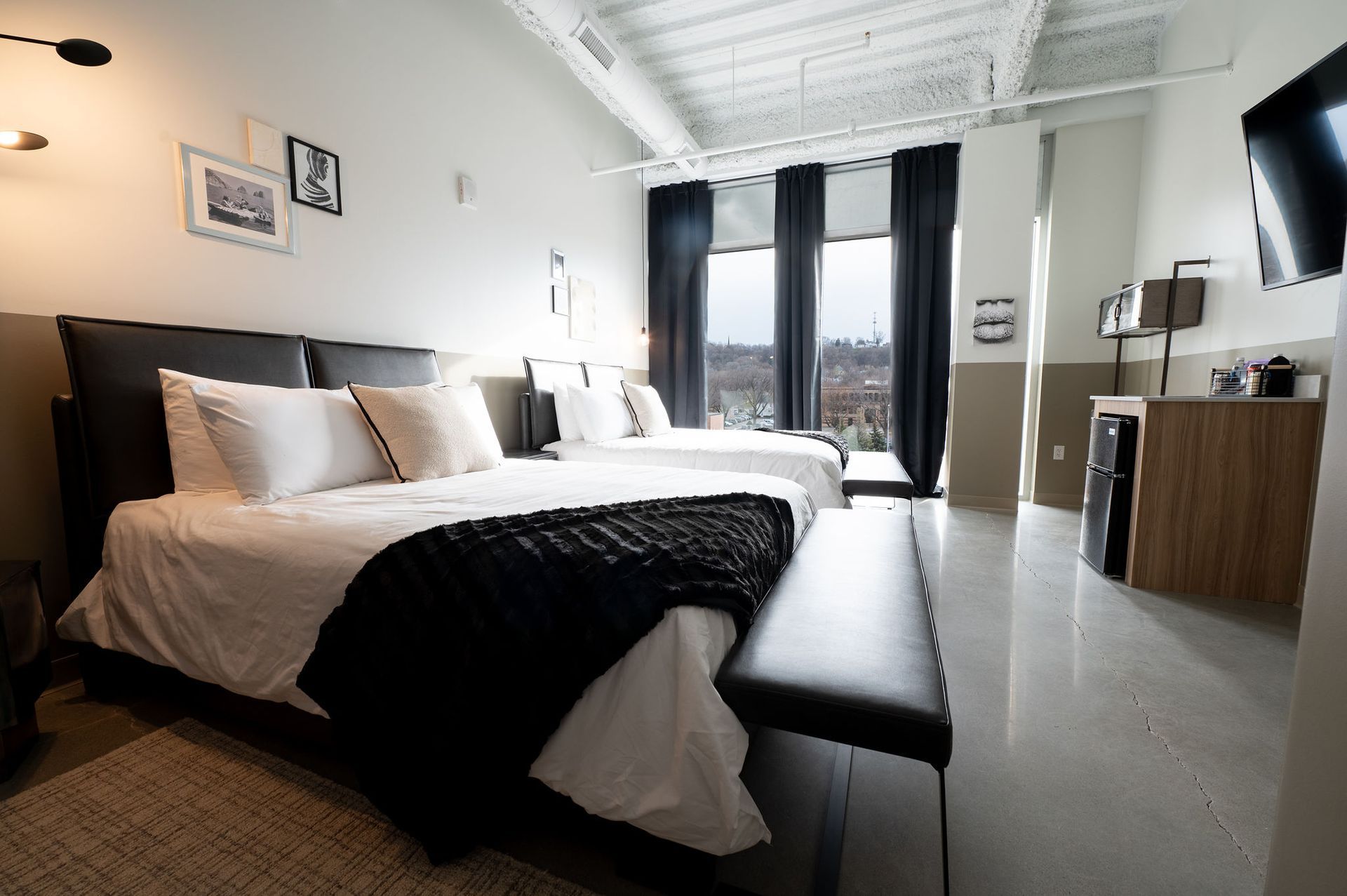 Modern bedroom with a neatly made bed, black bench, and large windows overlooking a city view