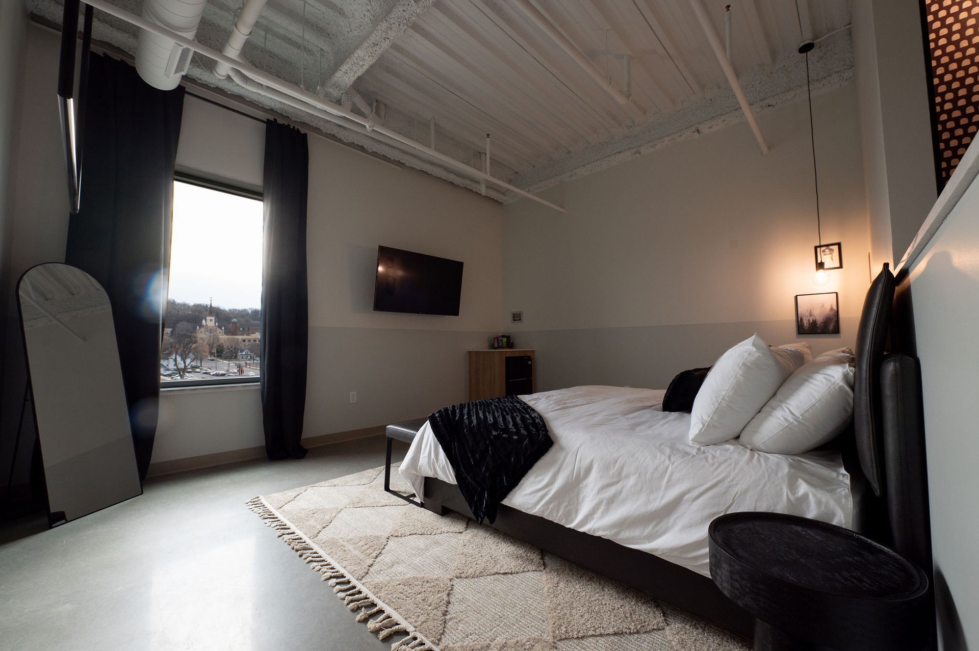 Modern bedroom with a large bed, wall-mounted TV, tall windows, and minimalist decor