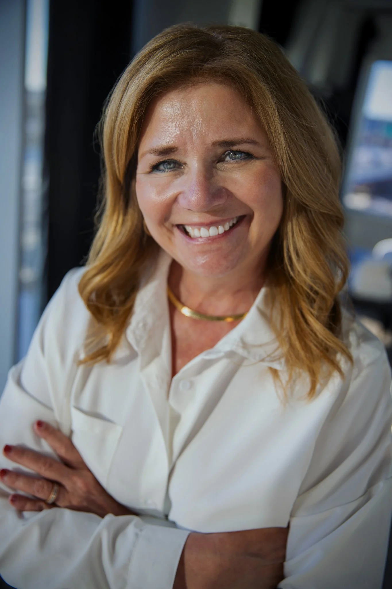 Smiling person in a white blouse with folded arms, standing indoors against a blurred background