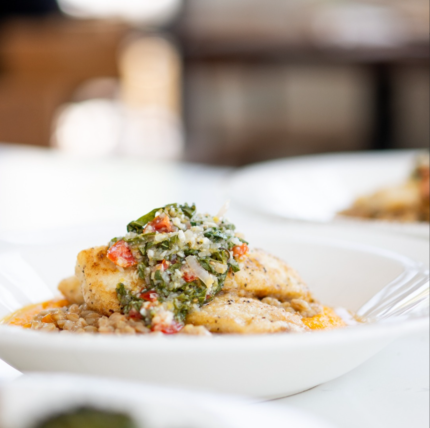 Plated chicken topped with herb salsa on a white dish in a softly blurred restaurant setting