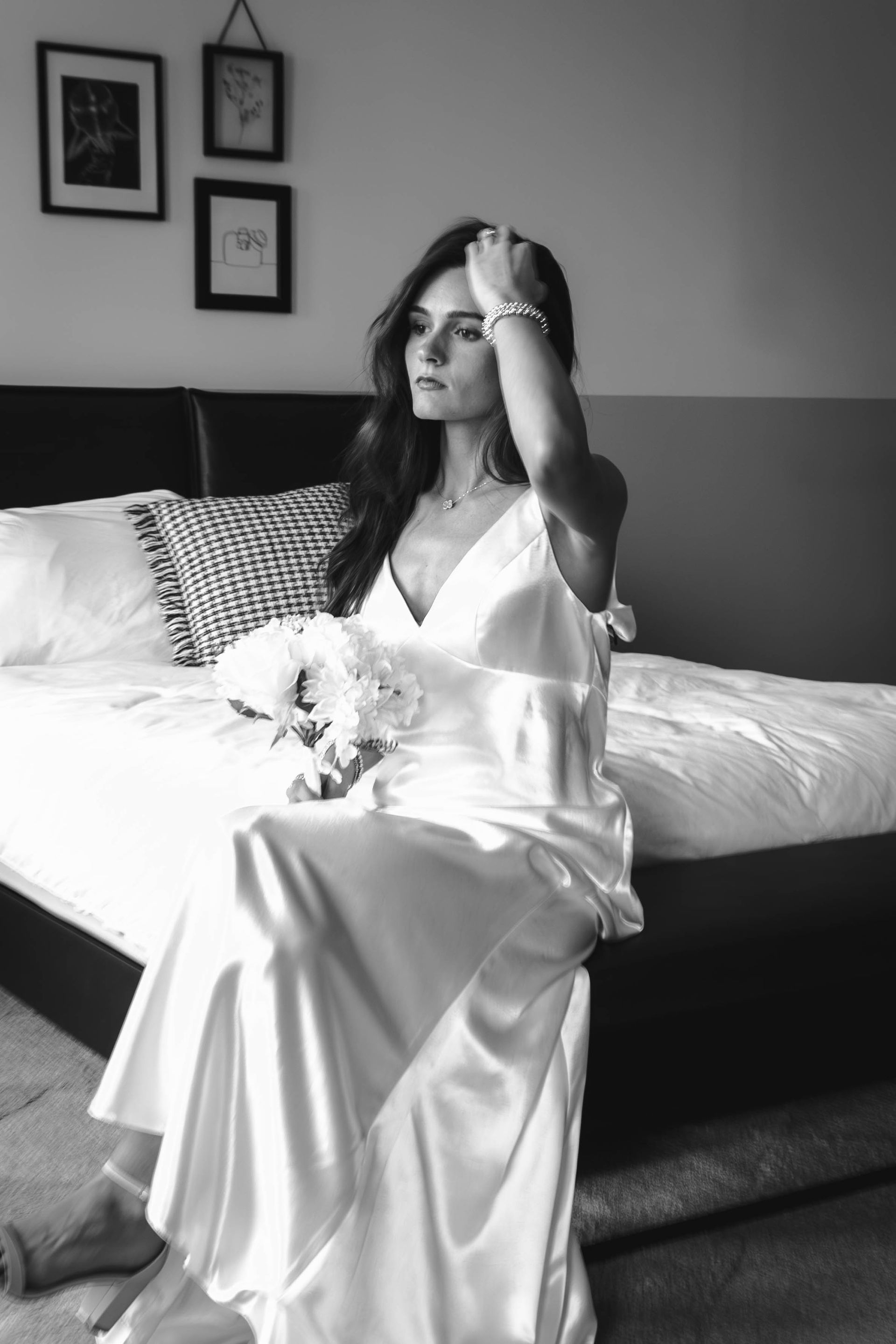 Woman in a satin dress sitting on a bed, holding flowers in a softly lit bedroom.