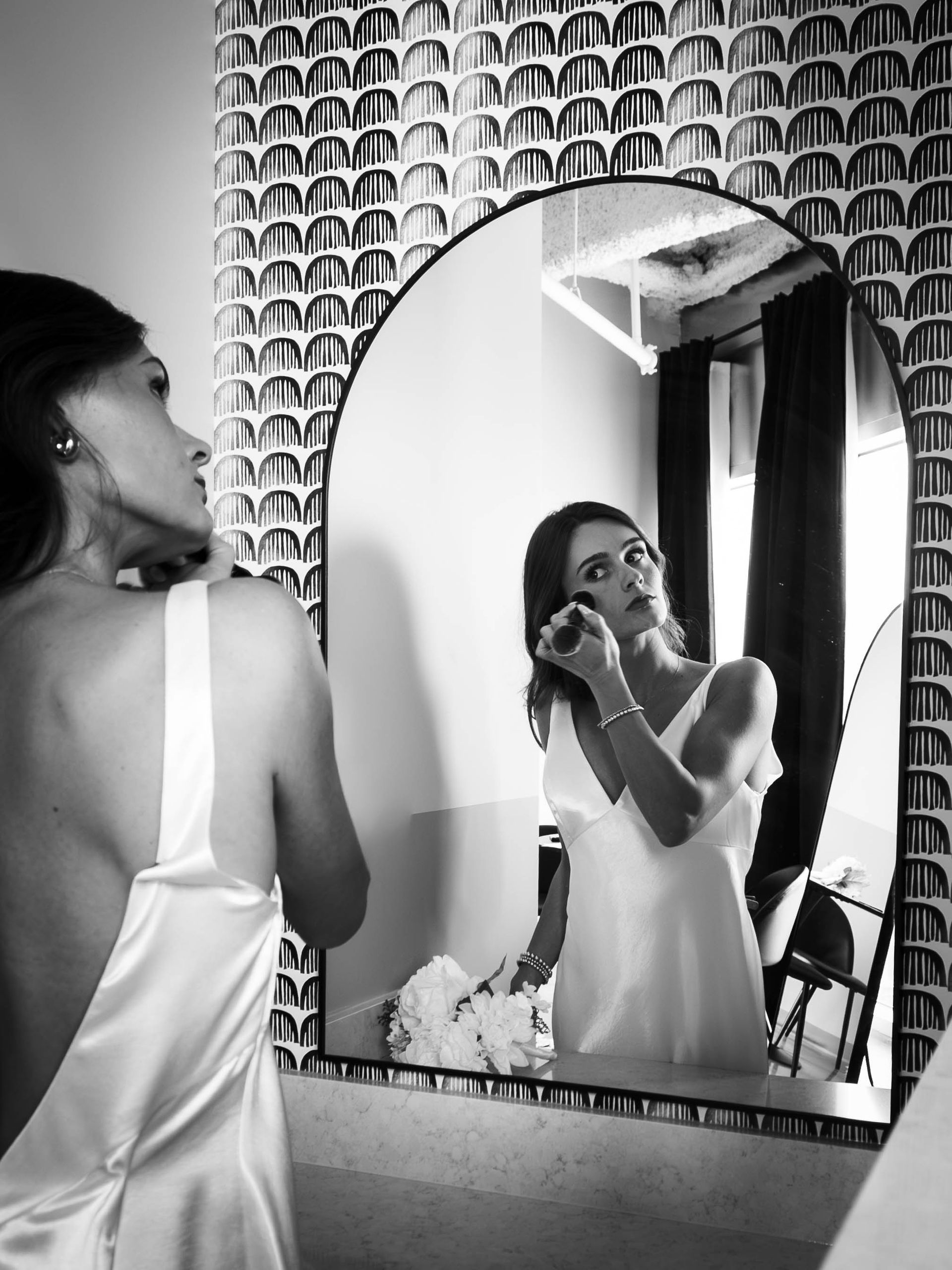 Woman in a white dress applying lipstick in a mirror, standing in a tiled bathroom.