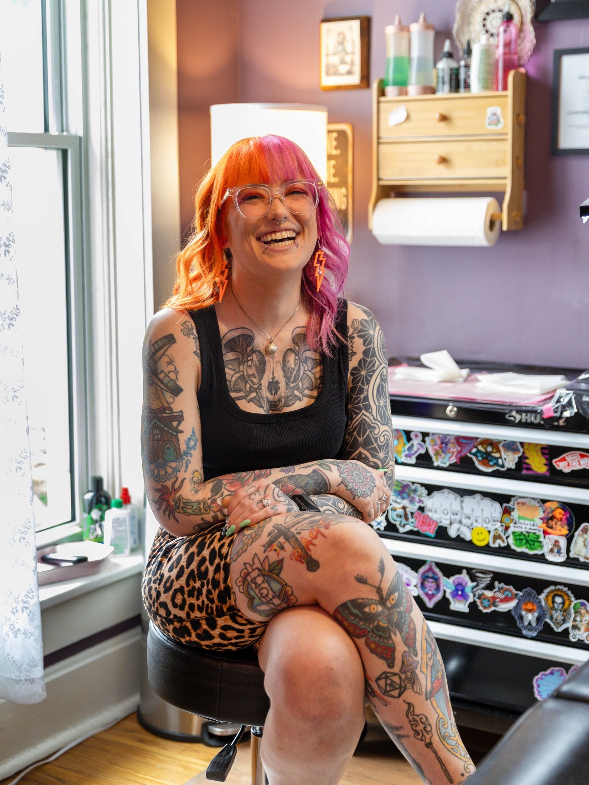 Woman with orange hair and tattoos sitting on a stool in a colorful room, smiling by a window.