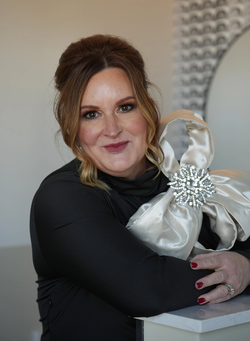 Woman in a black top holding a large silver satin bow, smiling indoors.
