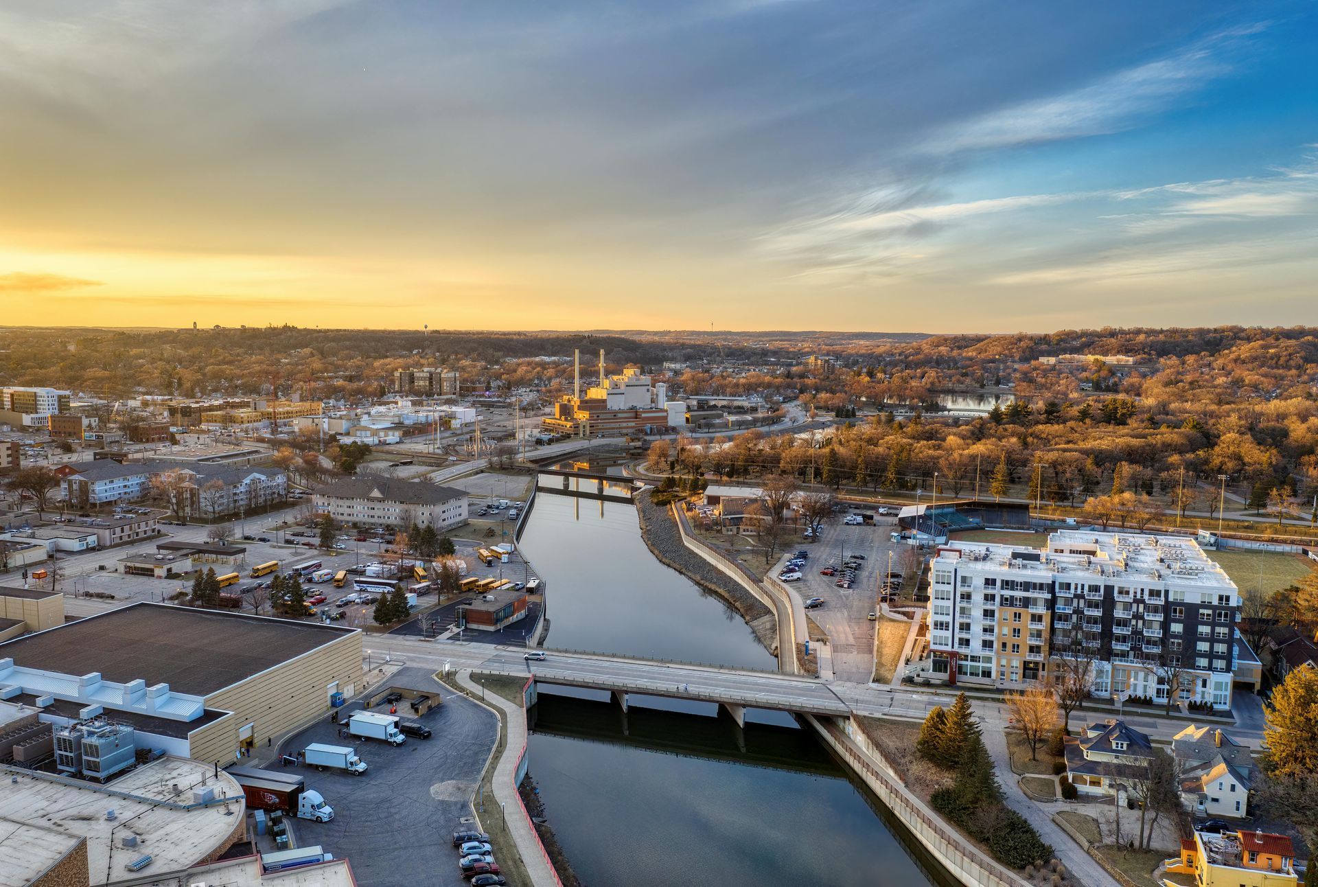 Snowy riverside city at sunset, with a bridge over the water and buildings lining the banks.