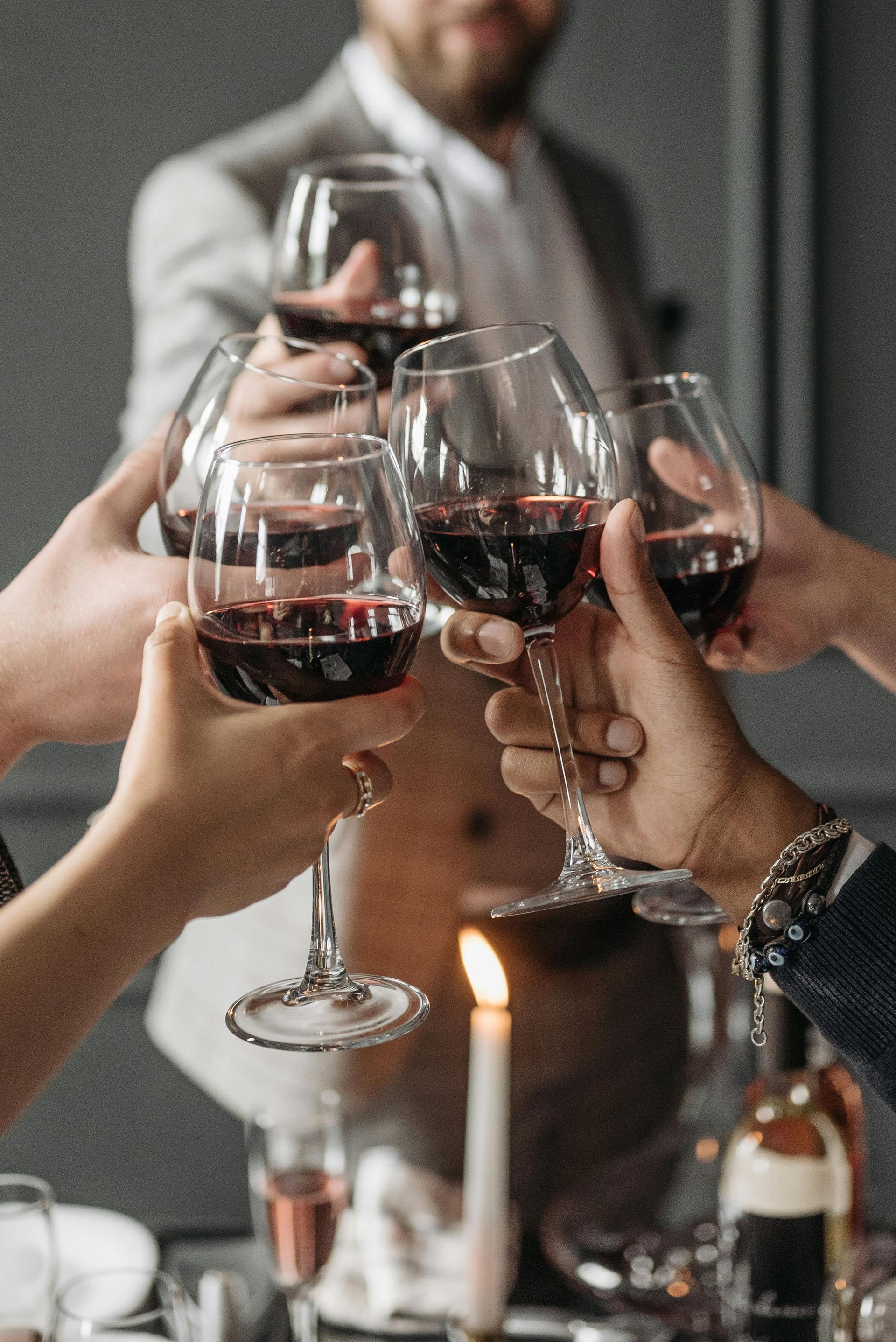 Hands toasting with red wine glasses around a candlelit table at an indoor gathering