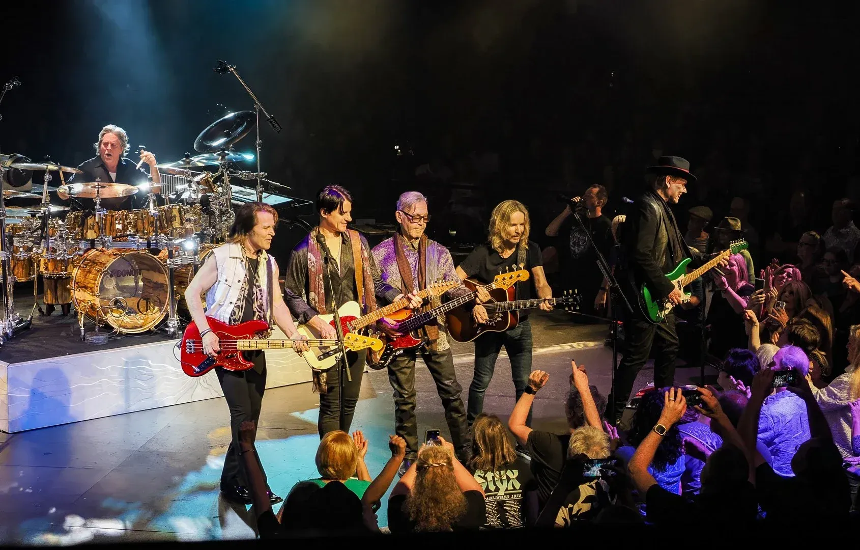 Rock band performing onstage under colorful lights, with drummer, guitarists, and cheering crowd in front
