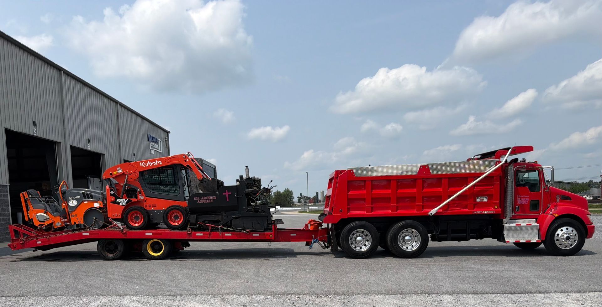 Red tow truck hauling a red dump truck trailer outside a warehouse under a cloudy sky