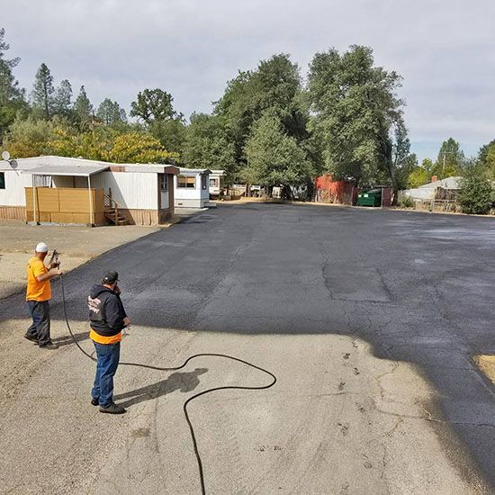 Two workers in orange vests spraying black asphalt in a parking lot with trees and trailers nearby