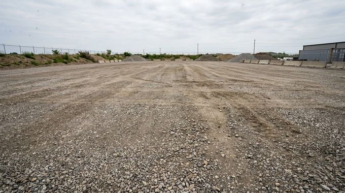 Empty gravel lot under an overcast sky, with sparse shrubs and buildings in the distance