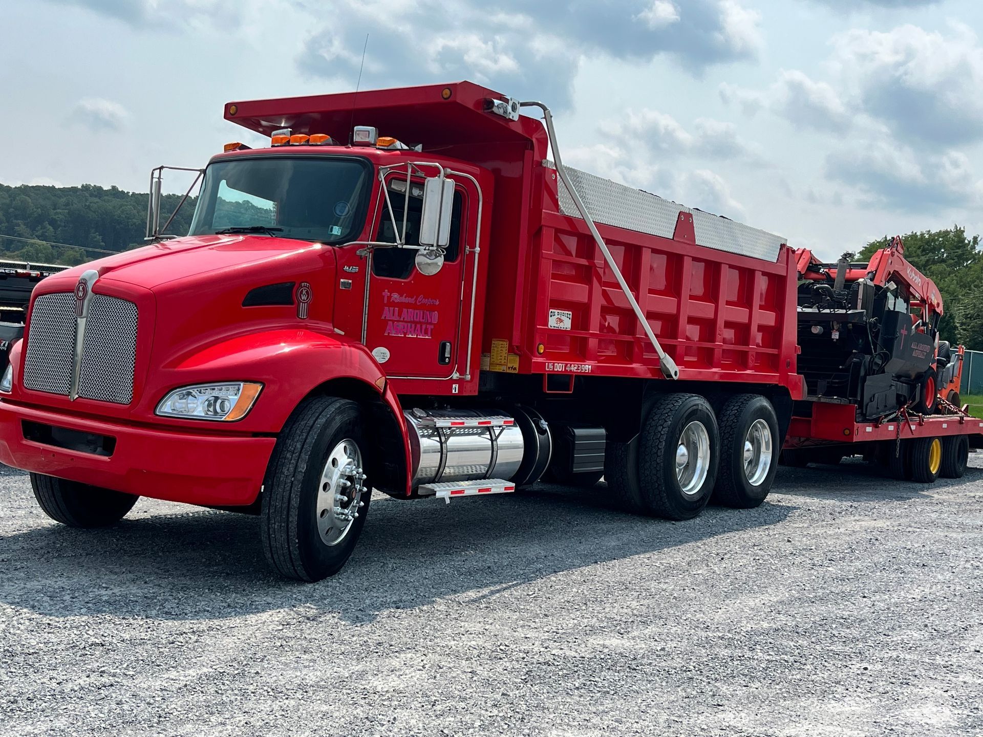 Red dump truck hauling heavy equipment on a gravel lot under a cloudy sky